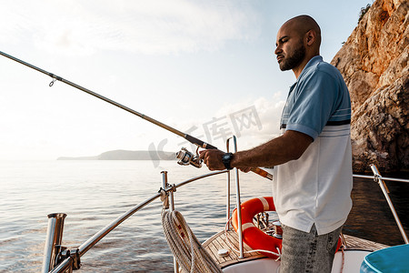 日落真实摄影照片_日落时，年轻的非洲裔美国人拿着钓鱼竿站在公海捕鱼的帆船上