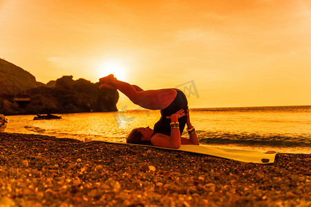 日落时，身穿黑色泳衣、戴着波西米亚风格手镯、长发的年轻女子在海边的瑜伽垫上练习户外。