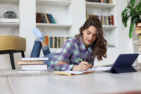 年轻的女学生在家学习，撰写要发表的文章，坐在地板上，在舒适的室内环境中使用平板电脑，周围是一堆书。