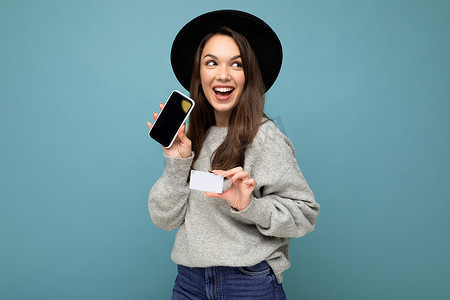 手持手机银行摄影照片_蓝色背景中，身穿黑色帽子和灰色毛衣的迷人、快乐的黑发年轻女性手持信用卡和手机的照片，显示空白，供模特看向侧面