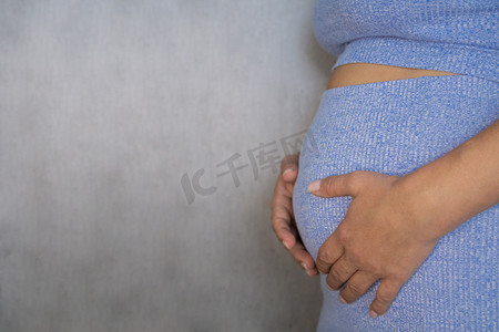 怀孕妈妈背景摄影照片_一名孕妇身着蓝色西装，在灰色背景上双手捧着肚子。