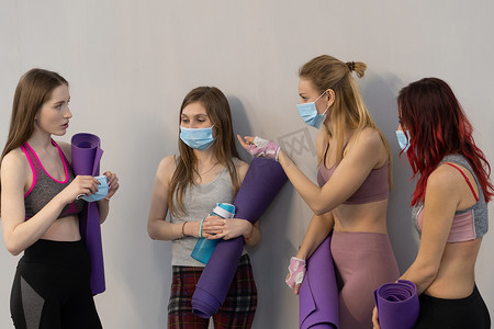 说服摄影照片_说服朋友戴上医用面罩，运动女孩穿着适合站在墙边拿着瑜伽垫，互相交谈，站在灰色背景下
