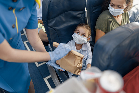 可爱的小杯子摄影照片_戴着防护面罩的可爱女孩看着空姐为机上的小乘客提供午餐。 