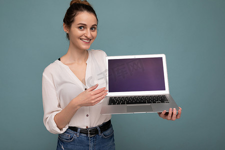 美丽快乐的年轻女子的特写肖像，她拿着笔记本电脑看着相机，开心地指着上网本显示屏，穿着休闲的智能衣服，与背景墙隔开