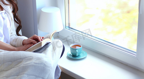 坐在椅子摄影照片_家里的年轻女人坐在窗边，在客厅里放松，看书，喝咖啡或茶。