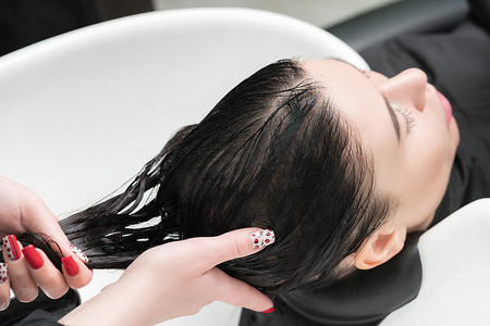 淋浴摄影照片_美发师的手用洗发专用水槽中的洗发水清洗黑发女性的长发