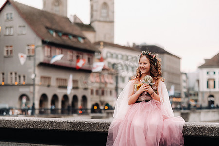 一个穿着粉红色公主裙的小女孩手里拿着一束花穿过苏黎世老城。瑞士城市街道上一个穿着粉红色连衣裙的女孩的肖像