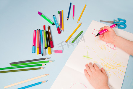 一个小孩用铅笔在相册、复制空间、顶视图、蓝色背景、铅笔、蜡笔、剪刀和桌子上的孩子的手上画画