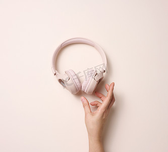 女性主义摄影照片_米色背景中的女性手持粉色无线耳机，顶视图