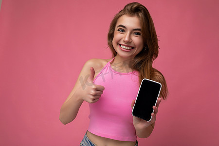 身穿粉红色上衣的美丽快乐的年轻金发女性，在粉红色背景中与世隔绝，手握空荡荡的空间，展示带有空显示屏的手机，用于模拟看着相机并竖起大拇指的手势