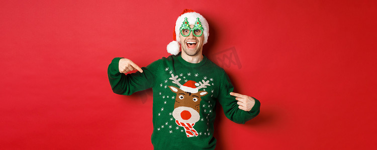 戴着派对眼镜和圣诞帽的快乐男人的形象，指着他的圣诞毛衣，微笑着，站在红色背景上