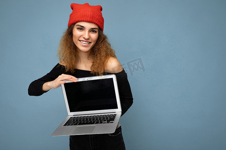 穿着黑色裁剪上衣和红色和橙色 do-rag 的美丽有趣微笑快乐的年轻黑发卷发年轻女学生隔离在浅蓝色墙壁背景上，拿着带有空屏幕监视器的电脑笔记本电脑看着相机