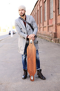 英俊的年轻人穿着灰色外套和帽子走在街上，使用长板。