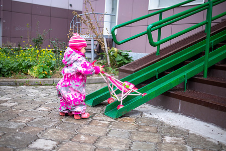 可爱设施摄影照片_带着玩具婴儿车的可爱蹒跚学步的孩子沿着轮椅、手推车和婴儿车的钢栏杆坡道行走。
