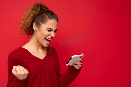 身穿深红色毛衣的年轻情绪化女性，与红色背景隔离，手持智能手机，玩得开心，看着手机屏幕，摆出肯定的手势