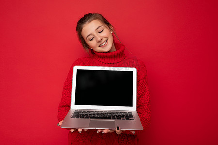 职业照摄影照片_照片中，美丽、满意、快乐的年轻女性手持笔记本电脑，低头看着上网本屏幕，身穿红墙背景中突显的红色毛衣