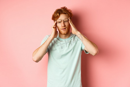 戴着弯曲眼镜的红发男子摸着头，感到头晕或恶心、宿醉或头痛，站在粉红色背景上