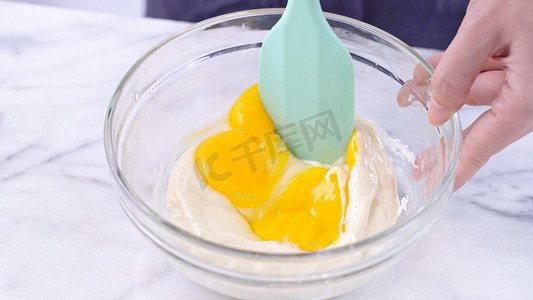蛋黄摄影照片_用绿色橡胶刮刀搅拌工具将蛋黄混合到蛋糕面糊中，搅拌至光滑，并在玻璃碗中充分混合，特写，生活方式