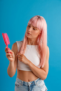 好奇的女人，一头光滑自然的粉红色长发，站在蓝色工作室背景中，一边看着梳子，一边与世隔绝