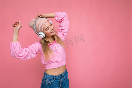 身穿粉红色衬衫和粉红色帽子的美丽快乐微笑的年轻金发女性，与粉红色背景墙上隔开，戴着白色无线蓝牙耳机，听着酷炫的音乐，玩得开心，跳舞