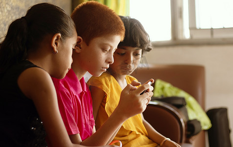 在家视频摄影照片_三个多民族的孩子或兄弟姐妹忙着在家玩手机游戏 — 儿童手机游戏成瘾的概念，使用技术、智能手机上的互联网。