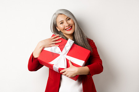 快乐的亚洲祖母抱着红色礼盒，微笑着感激，感谢礼物，站在白色背景上