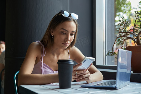 手机夏季夏季摄影照片_年轻的亚洲美女坐在咖啡馆的桌子旁，拿着笔记本电脑，使用智能手机