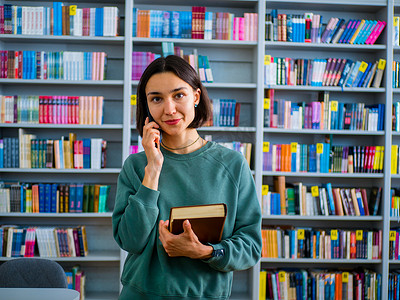 在书架的背景下，年轻的千禧一代女性站在公共图书馆里用手机交谈。