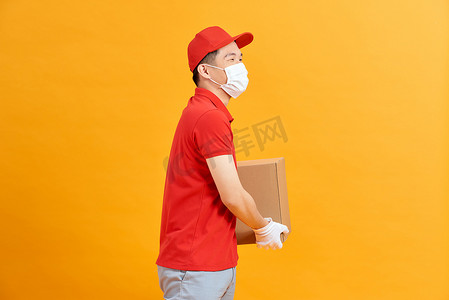 红帽摄影照片_身穿红帽空白 T 恤制服面罩手套的送货员拿着黄色背景中突显的纸箱