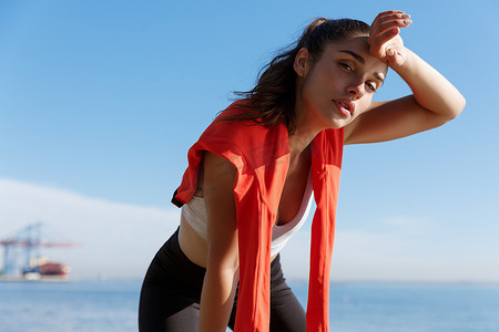 运动的人摄影照片_迷人的年轻健身女性在慢跑、喘息和擦额头上的汗水时休息的特写镜头，看着镜头很累，站在海边