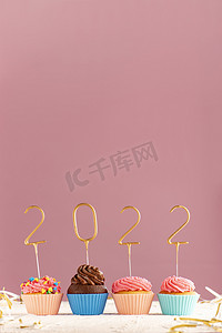 纸杯蛋糕或松饼，上面有奶油糖霜，粉红色背景上装饰着金色的 2022 年数字。