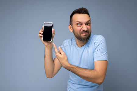 手机版k线摄影照片_愤怒英俊、胡子拉碴的黑发男子穿着日常蓝色 T 恤，与蓝色背景隔离，手持并展示手机，屏幕空空如也，可以看到相机并用手指指着智能手机