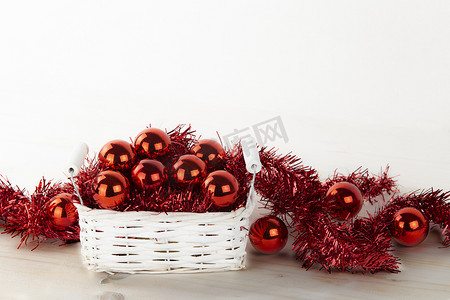 低角度圣诞复制空间：小破旧的白色篮子，里面装满了红色圣诞小饰品，浅色木桌和白色背景上有红色装饰花环