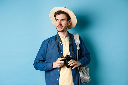 戴着夏日帽子的帅气正男，拿着相机，望向一边，游客在度假时拍照，背着背包站在蓝色背景上