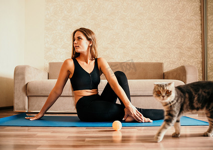 做运动的猫摄影照片_留着长头发的年轻女性，穿着黑色运动服的健身教练，在家里和猫一起在瑜伽垫上做伸展运动和普拉提。