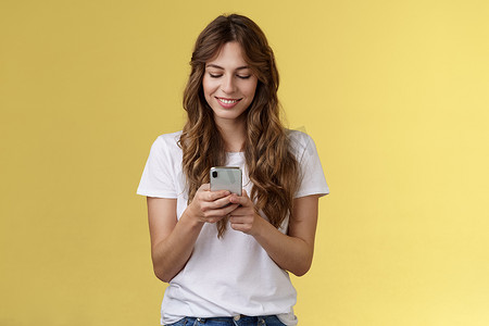 可爱的女性嫩白人女孩白色 T 恤牛仔裤拿着智能手机发短信的女朋友微笑着高兴的样子温柔可爱的微笑手机屏幕黄色背景