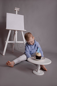 学龄前男孩穿着白色裤子和蓝色衬衫，灰色背景，靠近画架，上面有模型和带蜡烛的纸杯蛋糕，形状为四号 4。生日快乐。