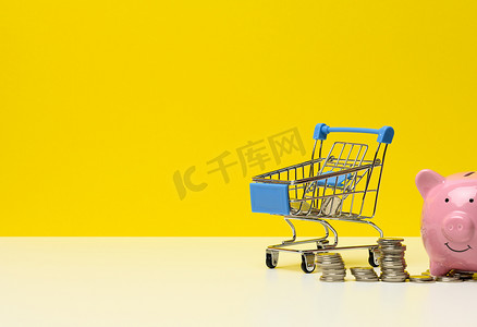 金属微型购物车，白色桌子上有零钱和粉色存钱罐，黄色背景。