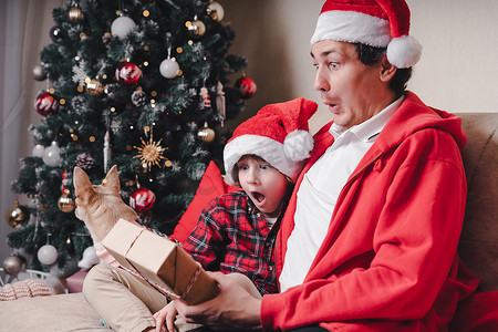戴着圣诞老人帽子的家人惊讶，父亲和孩子的儿子在家里打开圣诞礼物。