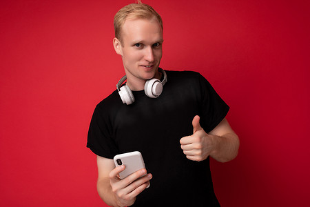 英俊帅气的金发年轻人身穿黑色 T 恤和白色耳机站在红色背景中孤立的照片，复制空间拿着智能手机，在手机上上网看着相机，竖起大拇指