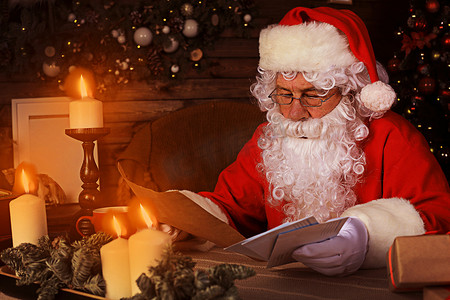 他在家摄影照片_快乐圣诞老人的画像坐在他在家里靠近圣诞树的房间里，阅读圣诞信或愿望清单。