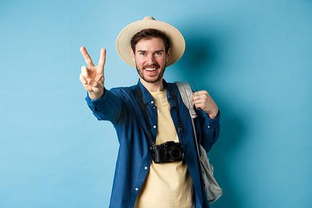 戴草帽的男摄影照片_帅气积极的男游客在暑假展示和平标志，在阳光下享受热带假期，戴着草帽，拿着背包和相机，蓝色背景