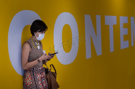 黄墙摄影照片_站在黄墙背景前，戴着口罩并使用智能手机打短信的年轻亚洲女性的侧视图。