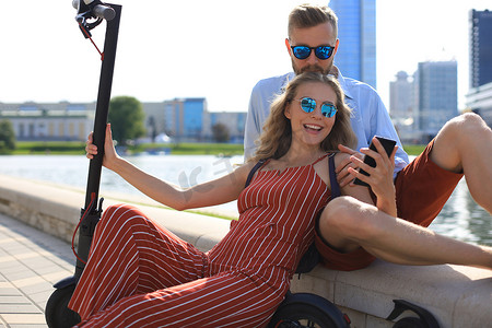 可爱的情侣开着电动滑板车玩得很开心，开车休息一下，坐在河岸上自拍