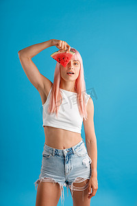 粉色丝滑丝滑摄影照片_可爱的年轻女人，一头粉红色的头发，眼睛上拿着一片西瓜，站在蓝色的工作室背景中，与世隔绝