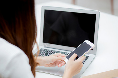 年轻女性的手在家里的办公桌上使用笔记本电脑和智能手机模型，笔记本和电话显示空白屏幕，自由职业者向互联网、商业和通信概念发送消息。