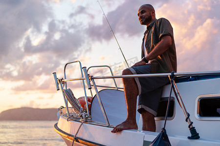 日落时，年轻的非洲裔美国人拿着钓鱼竿站在公海捕鱼的帆船上