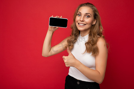 手握相机摄影照片_身穿红色背景中突显的白色 T 恤、面带微笑的美丽年轻金发女性，手握智能手机的复印空间显示手机，屏幕空空，用于模拟看着相机并竖起大拇指的手势