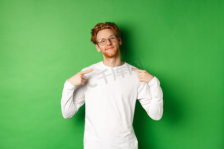 戴着眼镜、自信而自鸣得意的红发男子微笑着，自信地指着自己，站在绿色背景上