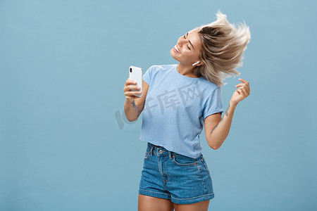 女孩在全新的无线耳机中享受酷炫的比特，通过智能手机在自己的博客录制视频中为耳塞做广告，在蓝色背景下欢快地跳舞，微笑着听音乐